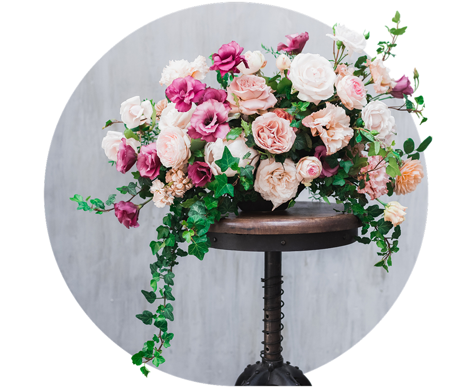 A floral arrangement with pink, white, and purple roses and green foliage sits on a round wooden stool against a light grey background.