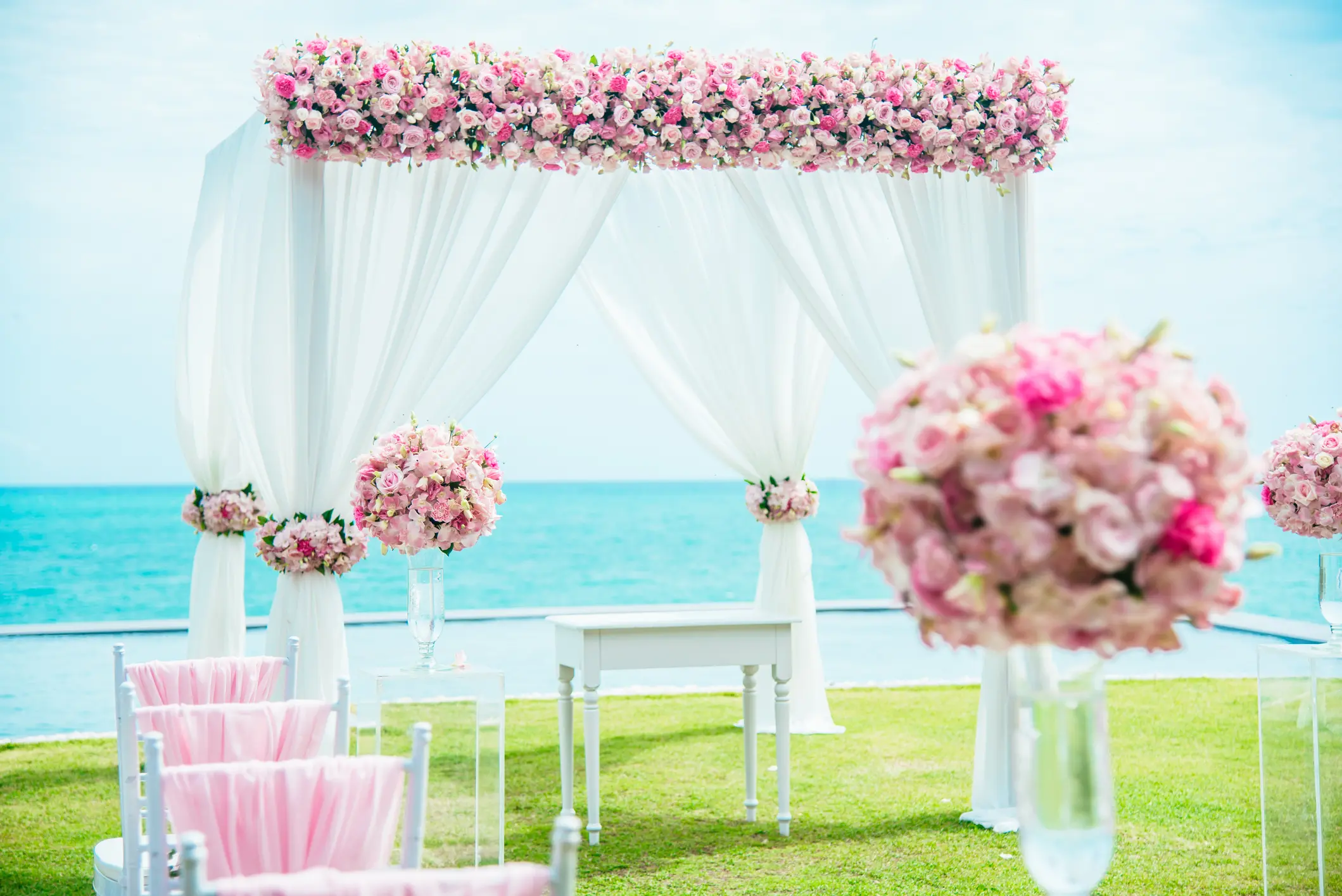 Outdoor wedding altar with white drapes and pink flower arrangements, set on grass with the ocean and sky in the background.
