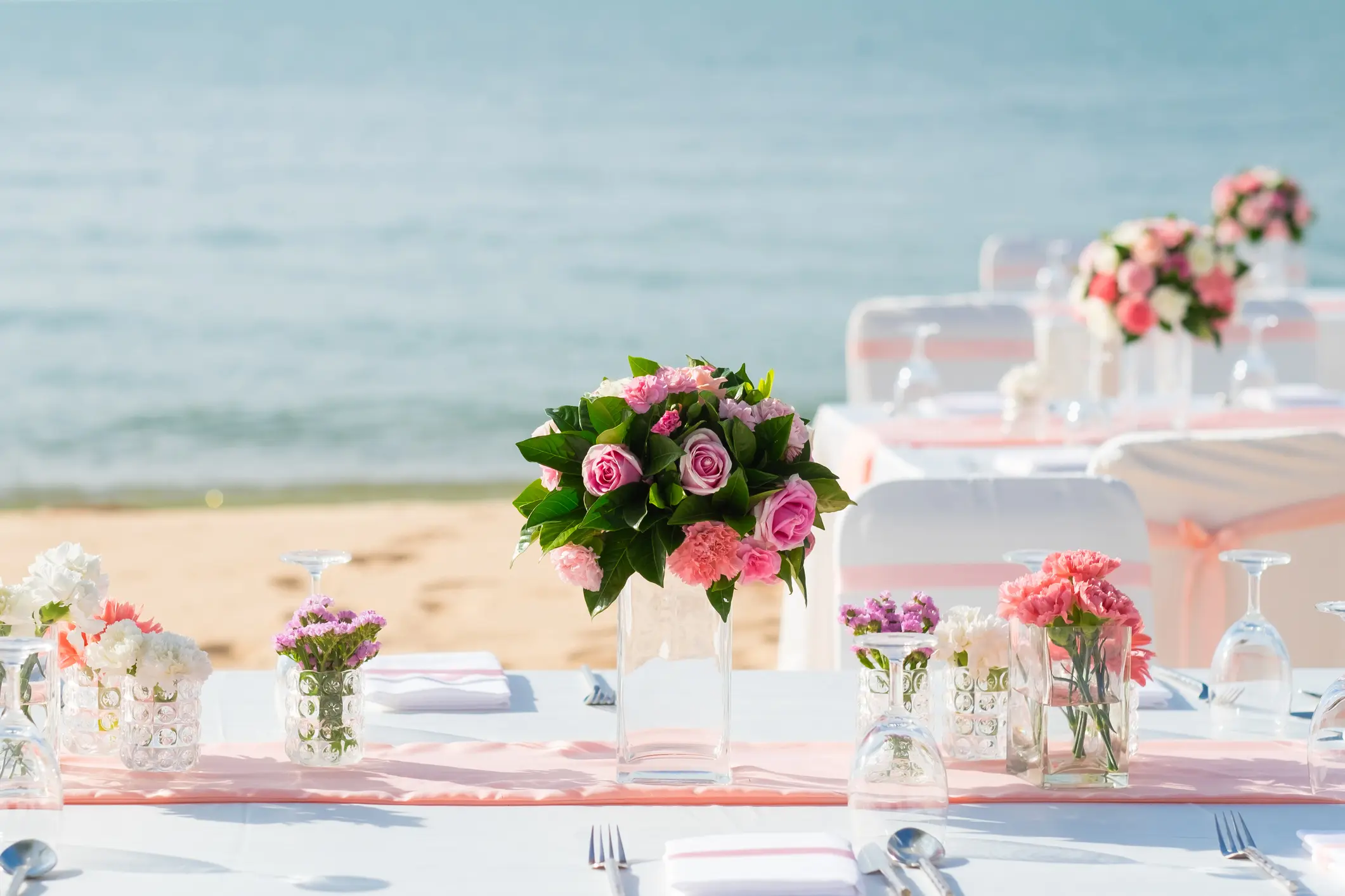 A decorated table set with pink and white flowers in glass vases is arranged outdoors on a beach with the sea in the background.