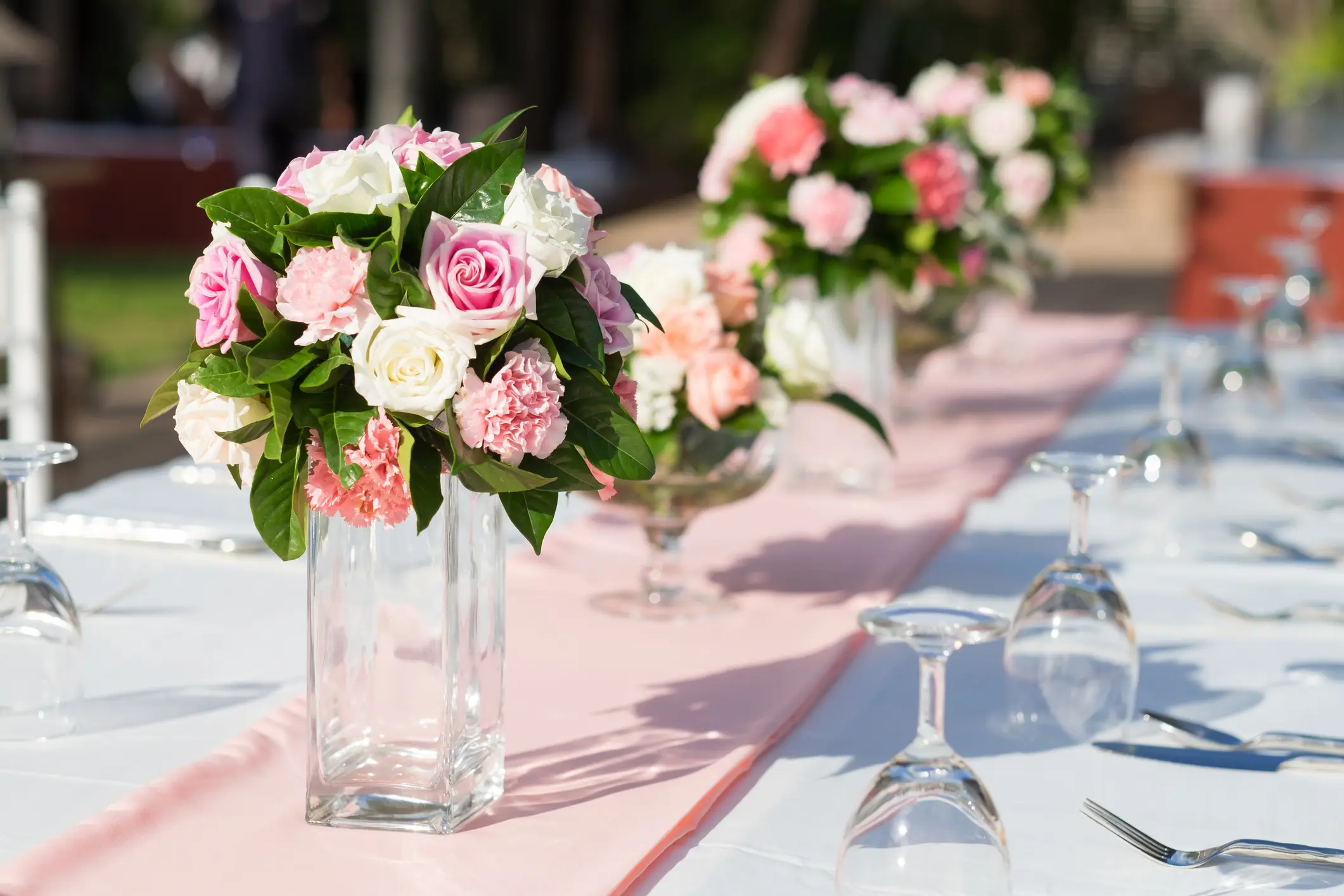 A close-up of a table set outdoors with glass vases of pink and white flowers, wine glasses turned upside down, and a pink table runner.