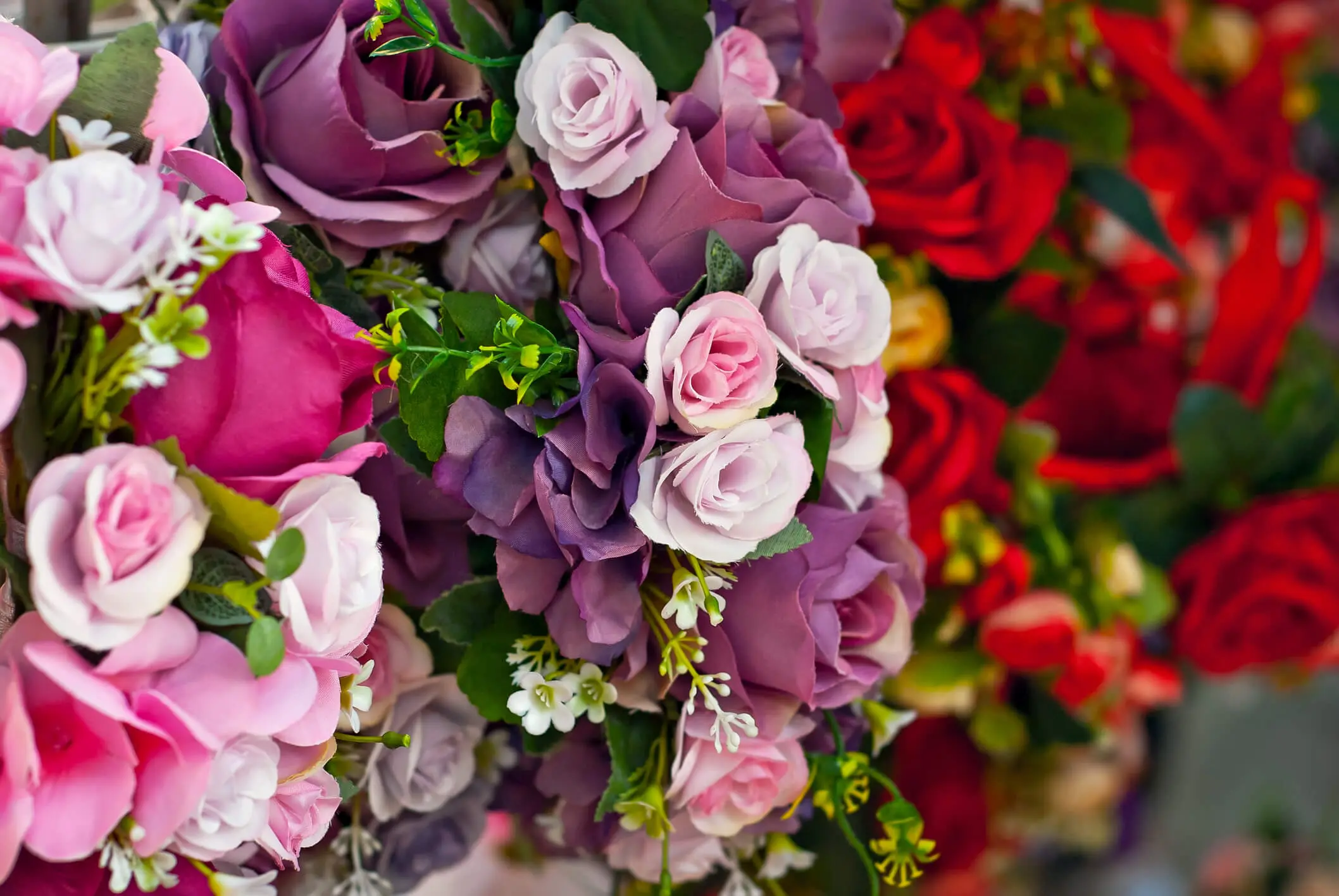 A close-up view of assorted artificial flowers in shades of purple, pink, and red, with green leaves and small white accents.