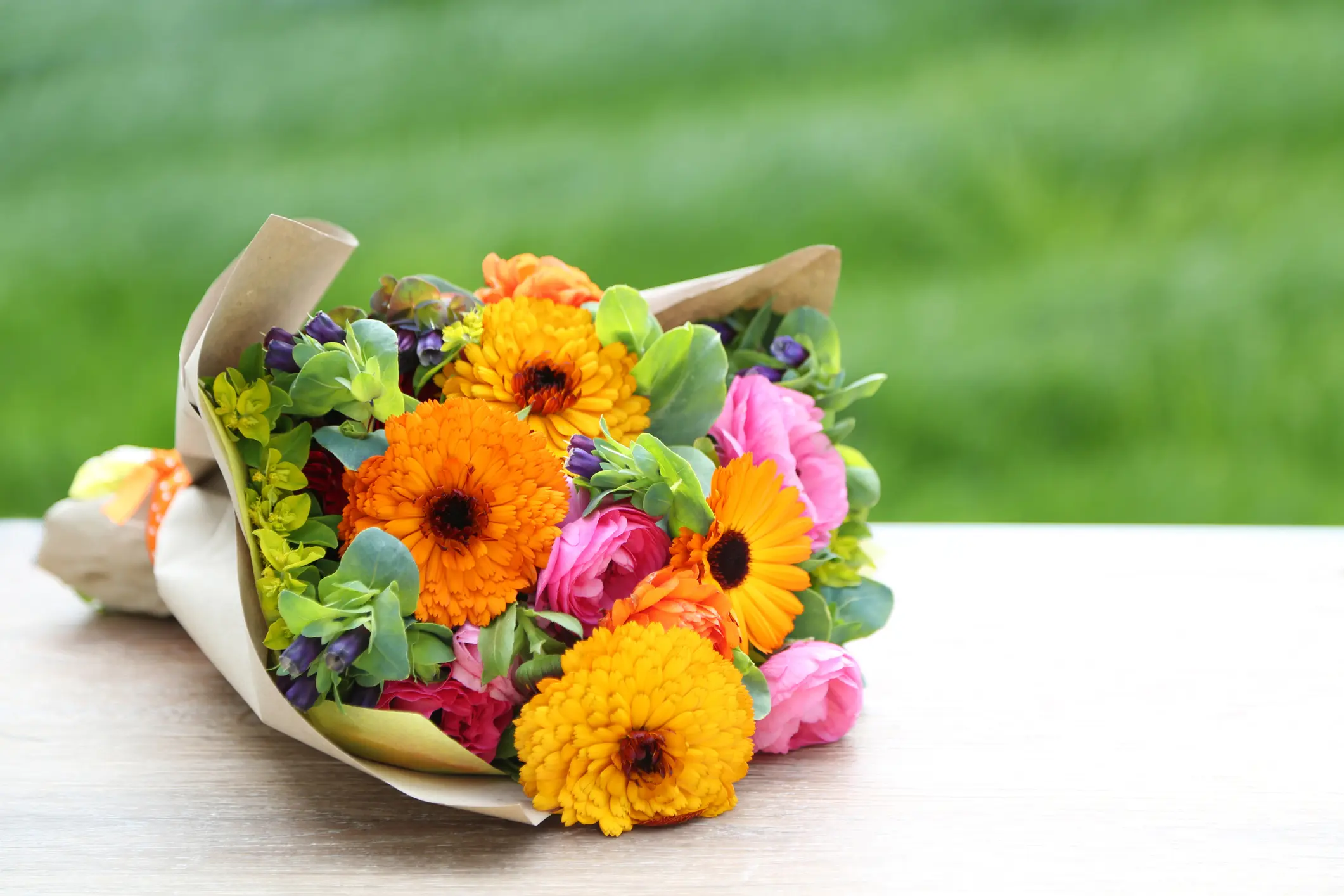 A bouquet of orange and yellow flowers mixed with green leaves and pink blossoms, wrapped in brown paper, resting on a wooden surface with a blurred grassy background.