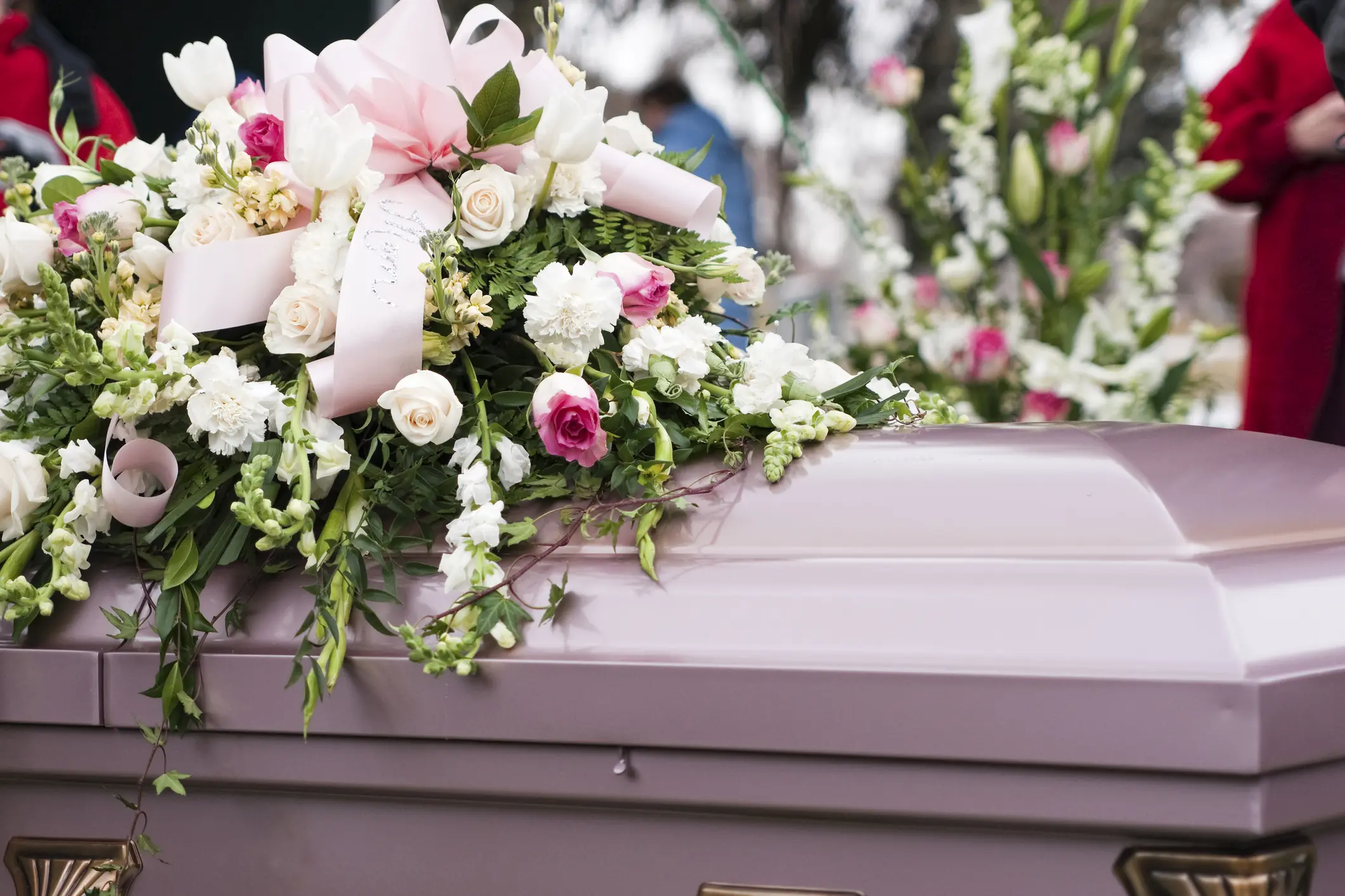 A closed, light purple casket is adorned with a large arrangement of white and pink flowers and a pale pink ribbon at a funeral.