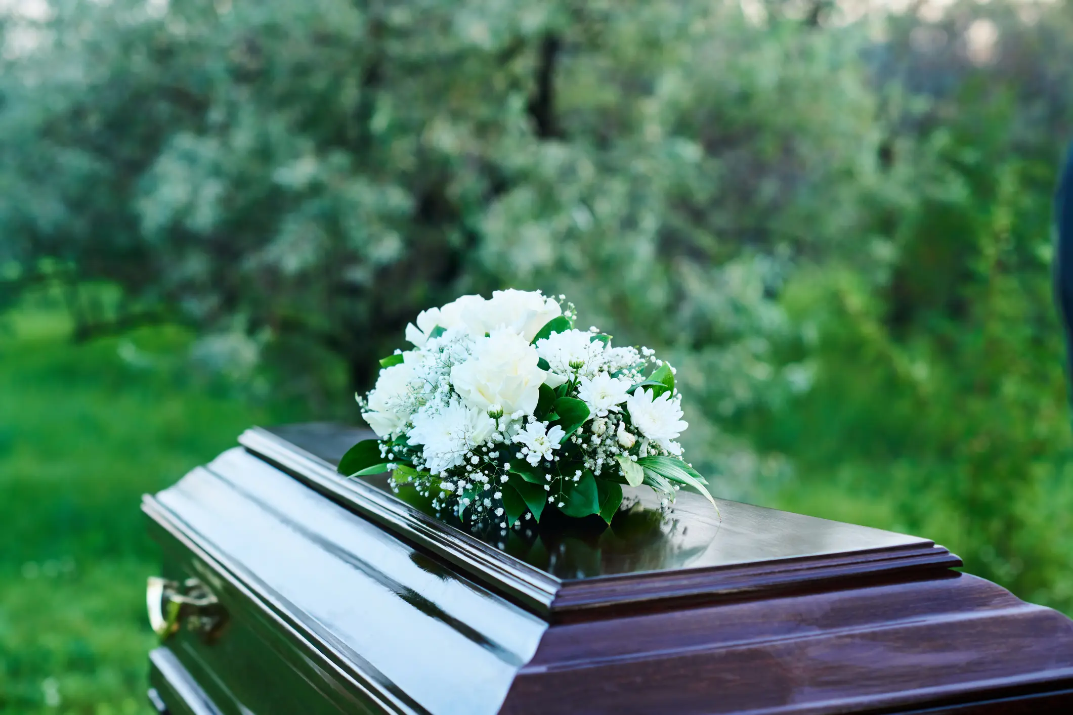 A wooden casket with a white floral arrangement rests outdoors in a green, grassy area.