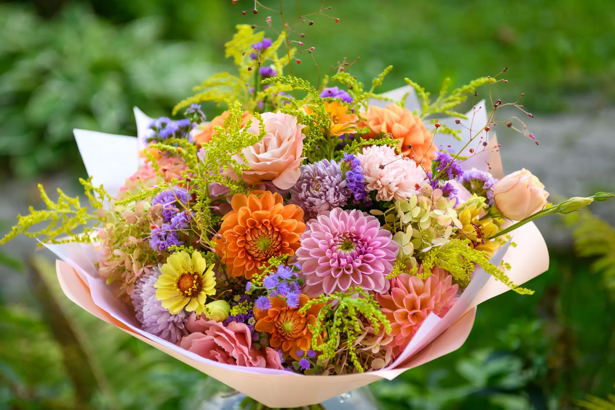 A bouquet of assorted flowers, including orange, pink, and yellow blooms, with greenery, wrapped in pale paper, set against an outdoor background.