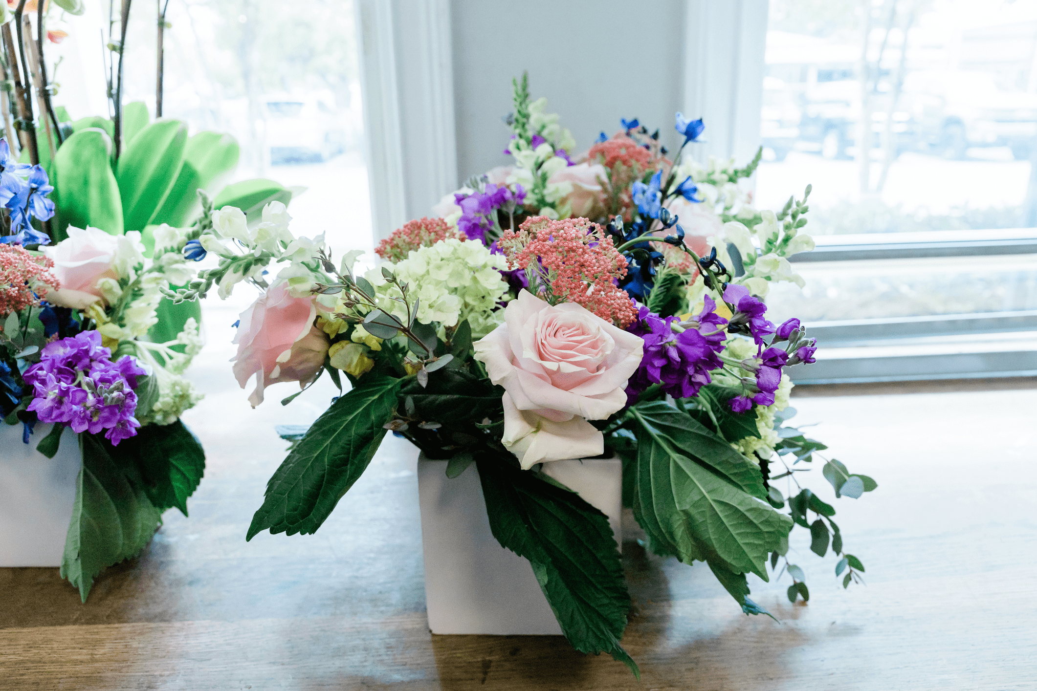 A rectangular white vase holds an arrangement of pink roses, purple and white flowers, and green leaves on a wooden table by a window.