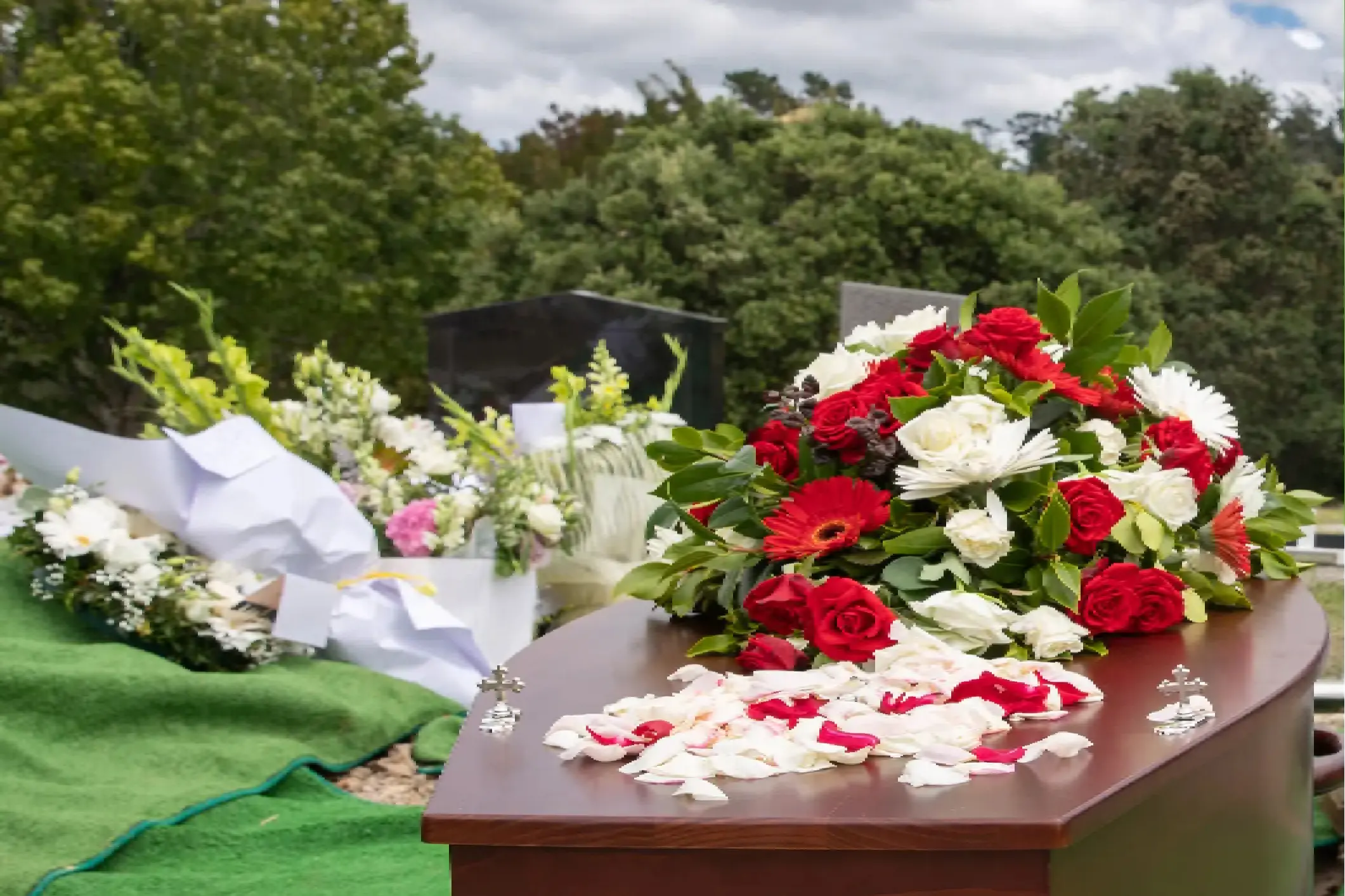 A casket with red and white flower arrangements and petals sits at a gravesite, surrounded by floral tributes on a grassy area under a cloudy sky.