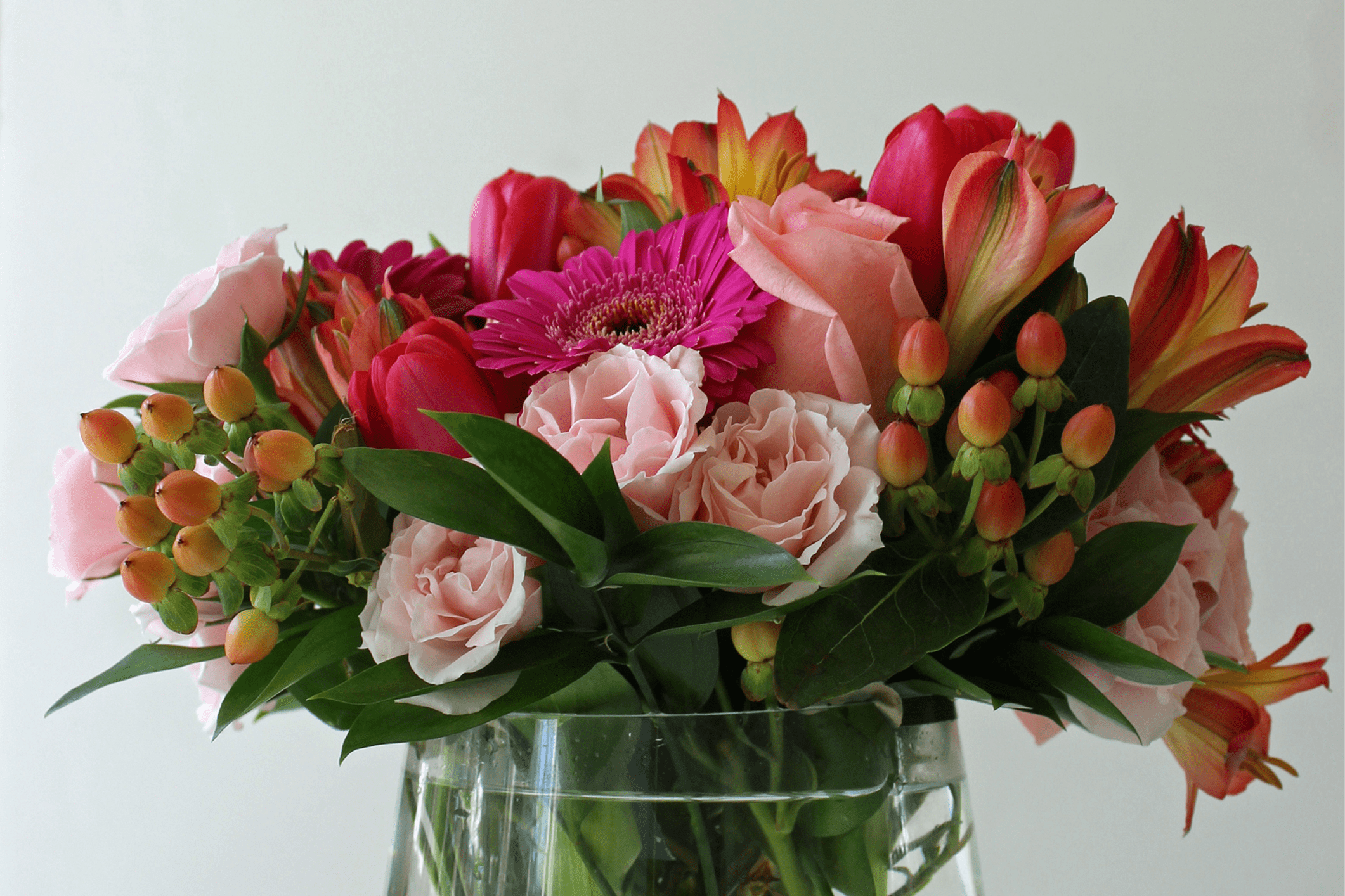 A clear glass vase filled with an arrangement of pink, red, and orange flowers with green leaves, set against a plain light background.