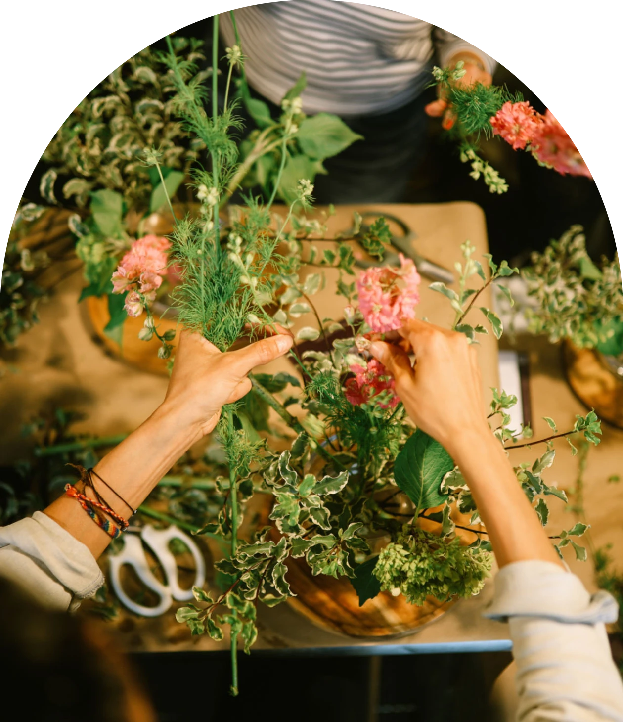 Person arranging a bouquet of flowers with various green leaves and pink blossoms on a table, with scissors visible nearby.