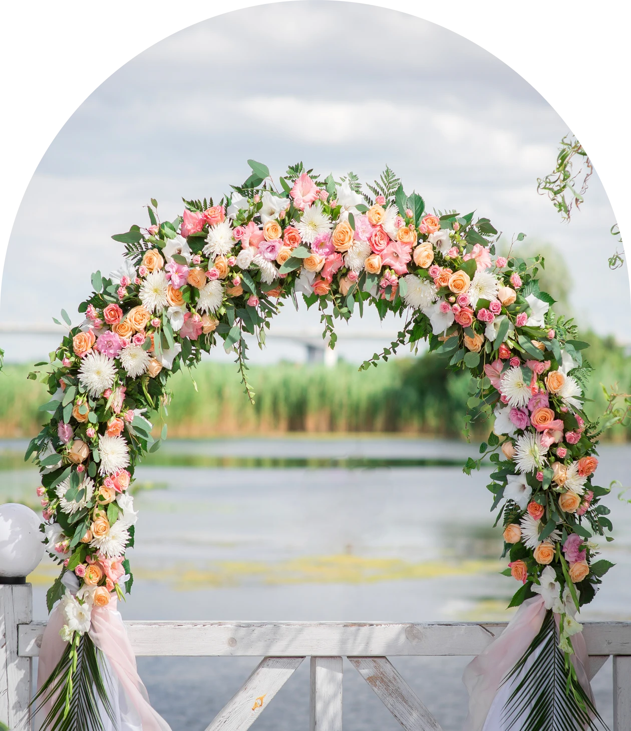 A floral wedding arch decorated with pink, white, and peach flowers stands outdoors by a lake on a cloudy day.
