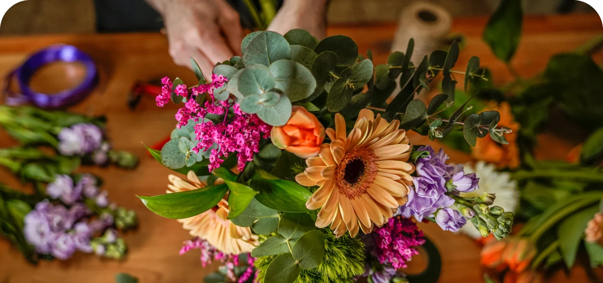 A person arranges a colorful bouquet of flowers, including gerbera daisies, eucalyptus, and various greens, on a wooden table with ribbon and twine nearby.