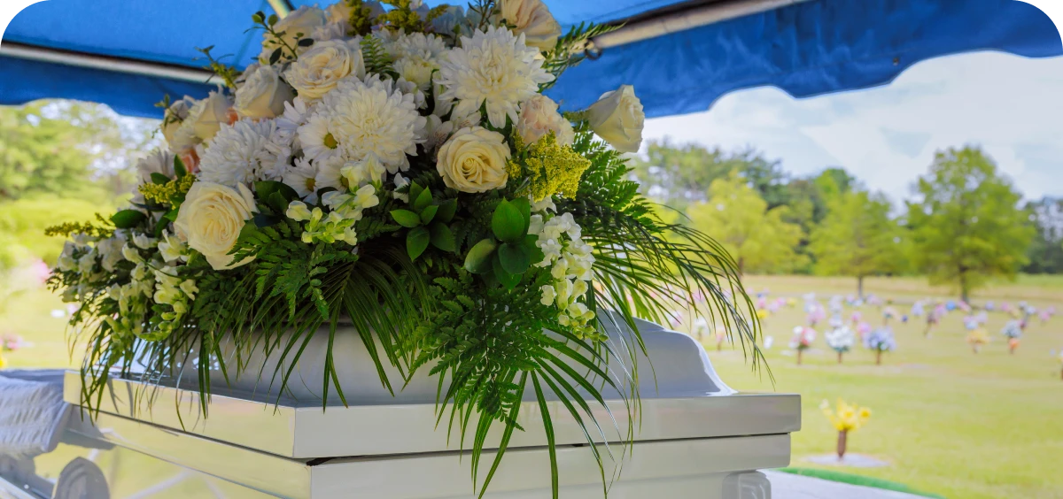 A white coffin with a floral arrangement of white and yellow flowers sits outdoors under a blue canopy at a cemetery.