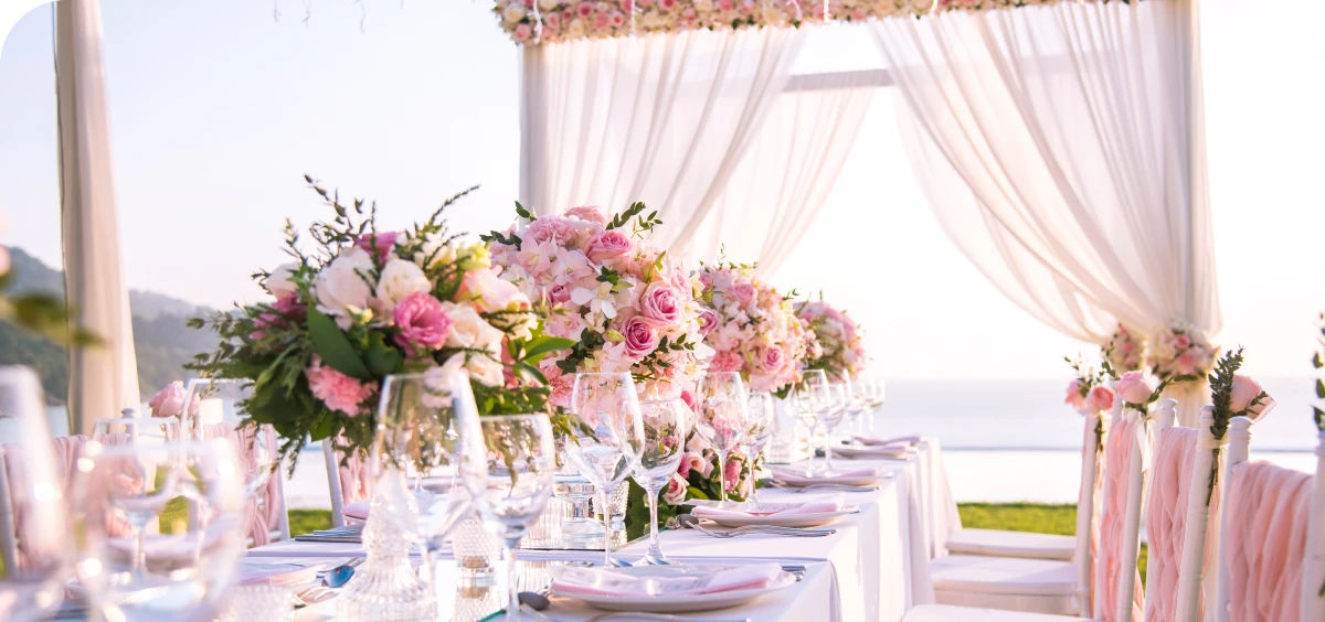 Elegant outdoor wedding table setup with pink and white floral arrangements, glassware, and draped white fabric canopy overlooking a scenic landscape.